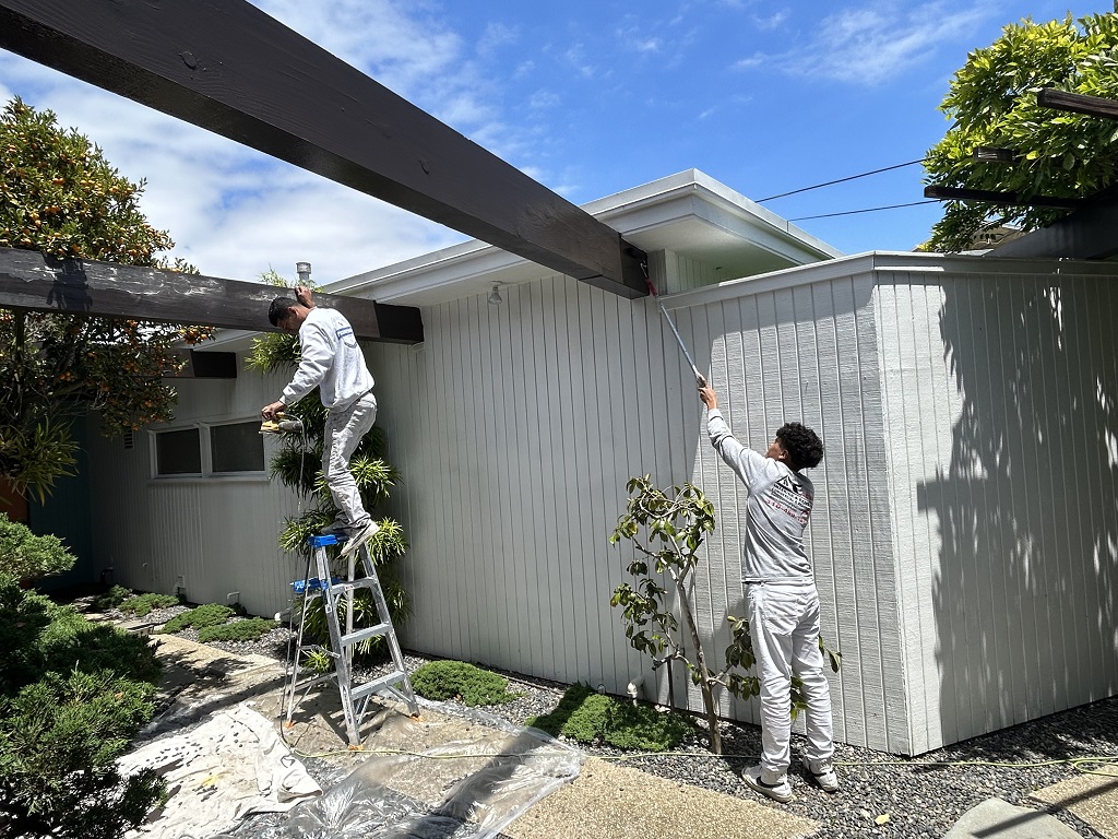 an image of two workers painting 3 horizontal woods attached to the house with black paint